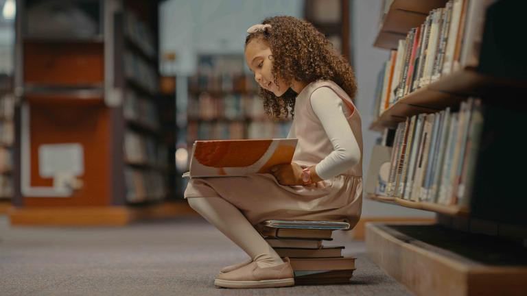 Child sitting on a stack of books reading a book in a library.